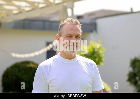 Ein reifer Mann in einem weißen T-Shirt lächelt sanft, während er in einem sonnendurchfluteten Garten steht. Stockfoto