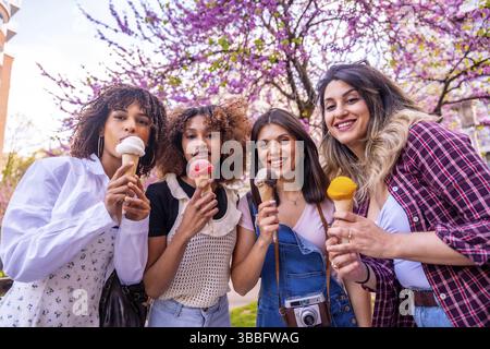 Vier verschiedene Freundinnen genießen gemeinsam an einem sonnigen Frühlingstag unter wunderschönen blühenden Bäumen Eiskegel Stockfoto
