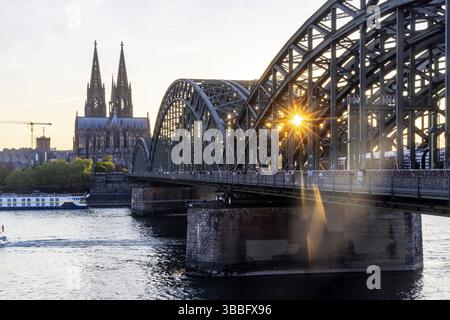 Kölner Dom am Abend mit Hohenzollernbrücke, Eisenbahnbrücke über den Rhein. Sonnenuntergang auf dem Rhein mit Sonnenstern. Stadtblick auf Köln, NOR Stockfoto