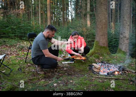 In einem üppigen Wald bereitet ein Paar eine Mahlzeit über einem offenen Feuer zu, eingetaucht in die Klänge der Natur, während die goldene Stunde ein warmes Licht auf ihre Outdoor-Kochen wirft Stockfoto