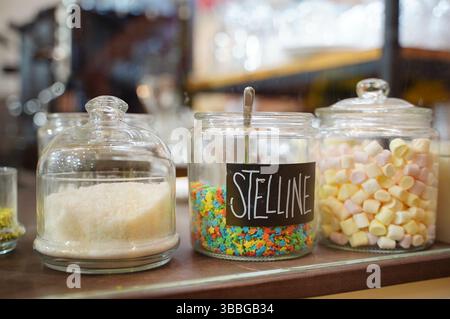Gläser mit farbenfrohen Dekorationen und Marshmallows werden auf einer Arbeitsplatte in einem Café ausgestellt. Das mittlere Gefäß ist mit Stelline beschriftet Stockfoto