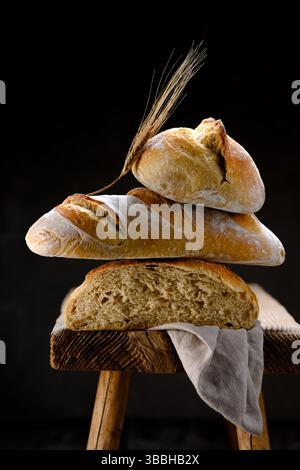 Komposition aus knusprigem Sauerteig-Baguette, Brotbrot und Brötchen auf einem rustikalen Holzhocker mit Ohr und Leinentuch. Handwerklich anmutende Loafs im Bauernhausstil in warmer natürlicher Beleuchtung Stockfoto