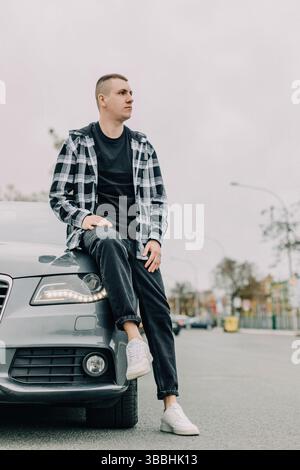 Junger Mann in urbanem Outfit, der sich auf der geparkten Motorhaube ausgibt Stockfoto