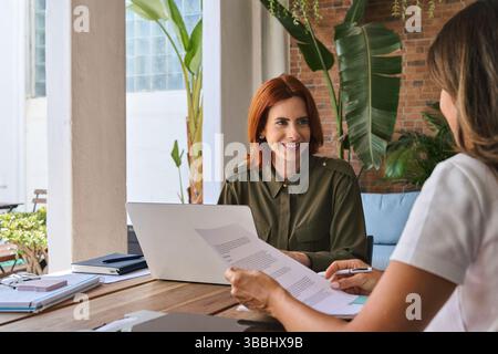Junge glückliche Managerin berät Klientin, die im Büro mit Laptop arbeitet. Stockfoto