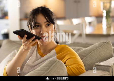Das Smartphone in der Nähe des Mundes hält, Asiatin in ihren Zwanzigern sitzt auf dem Sofa im Wohnzimmer Stockfoto
