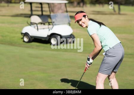 Lächelnde Frau mit Korallenvisier, die Putter in der Nähe von Golfball auf Putting Green hält, Kopierraum Stockfoto