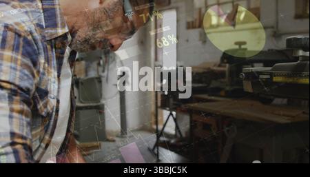 Anlehnender Zimmermann, der Holzstücke an der Werkstattwerkbank bearbeitet, mit Kapselgehörschützern und Tischsäge Stockfoto