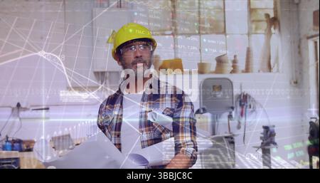 Mann mit gelbem Schutzhelm und Brille, der Pläne in der Werkstatt hält, mit überlagerten Finanzdiagrammen Stockfoto