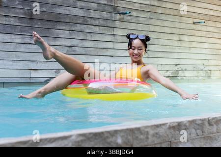 Entspannende asiatische Frau in orangefarbenen Badeanzügen mit Sonnenbrille auf aufblasbarem Ring im Pool zurückgelehnt Stockfoto