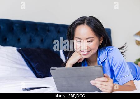 Lächelnde asiatische Frau mit blauem Satin-Pyjama, die im Schlafzimmer auf dem Bett liegt, mit Tablet, Kopierraum Stockfoto