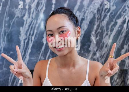 Asiatische Frauen blinkende Friedensschilder im Spa-Badezimmer mit rosafarbenen Augenflecken und Goldschmuck Stockfoto