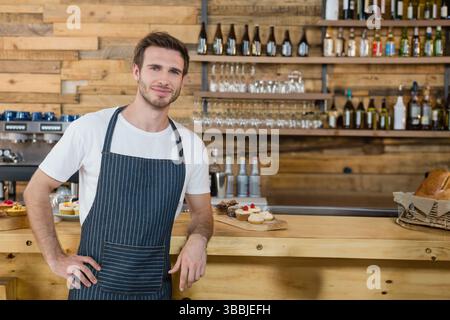 Lächelnder männlicher Barista, der sich auf eine rustikale Holztheke im Café lehnt und verschiedene Gebäckstücke und Brot präsentiert Stockfoto