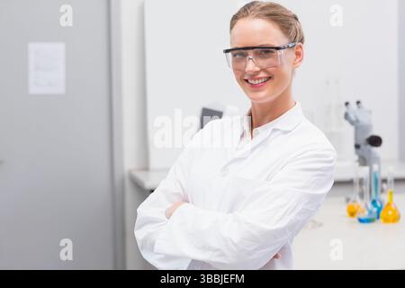Stehende Wissenschaftlerin mit Laborkittel und überkreuzten Armen im Labor, mit Mikroskop und Kolben Stockfoto