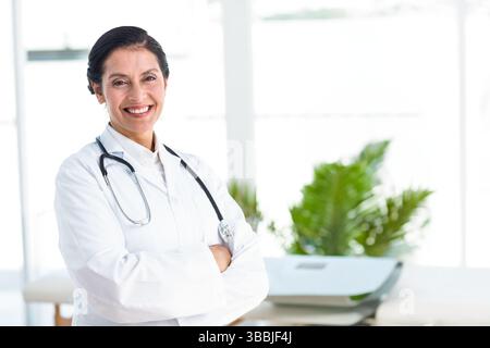 Lächelnde Ärztin, die Arme kreuzt, während sie in der Klinik steht, mit Stethoskop, Kopierraum Stockfoto