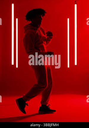 Tanzender afroamerikanischer Mann mit Hoodie und Jogginghose in rotem Studio, mit vier LED-Röhrenlichtern Stockfoto
