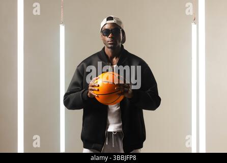 Posierender afroamerikanischer Mann mit Jacke, der Basketball im Studio hält, mit LED-Röhren Stockfoto