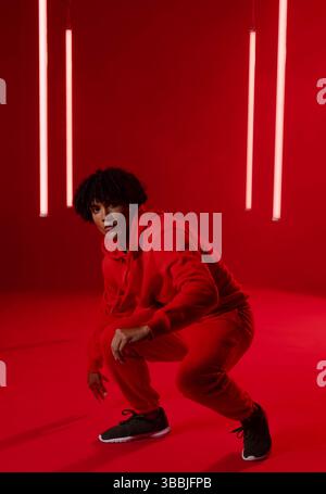 Hockender Afroamerikaner mit rotem Sweatsuit in rotem Studio, vertikale LED-Leuchtröhren Stockfoto