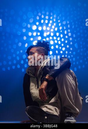 Ein Mann in der Hocke Ende 20, der sich im Studio auf das Knie gelehnt hat, mit Lederjacke, Hoodie, Sneakers Stockfoto