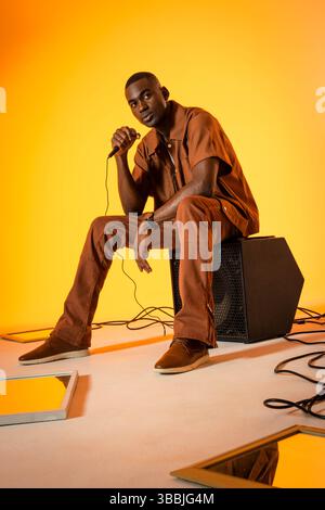 Sitzender afroamerikanischer Mann mit Mikrofon auf Lautsprecher im Studio, mit Kabeln und Spiegeln Stockfoto
