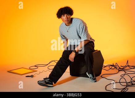 Sitzender Mann mit blauem Pullover und Turnschuhen auf Lautsprecher im Studio, mit Mikrofon, verwickelten Kabeln Stockfoto