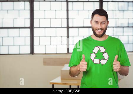 Lächelnder Mann mit grünem Recycling-T-Shirt, der am Arbeitsplatz mit Tisch und Kopierraum die Daumen nach oben zeigt Stockfoto