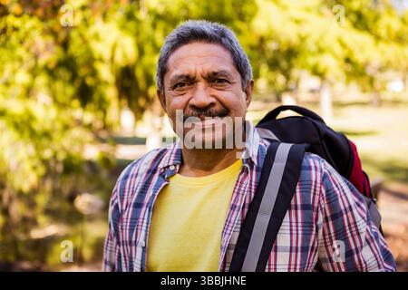 Lächelnder älterer Mann, der auf dem Weg im sonnendurchfluteten Park spaziert, mit rot-schwarzem Rucksack auf der Schulter Stockfoto