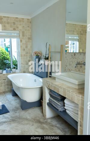Die freistehende weiße Badewanne befindet sich im modernen Badezimmer mit einer Holzleiter mit rosafarbenen Blumen Stockfoto