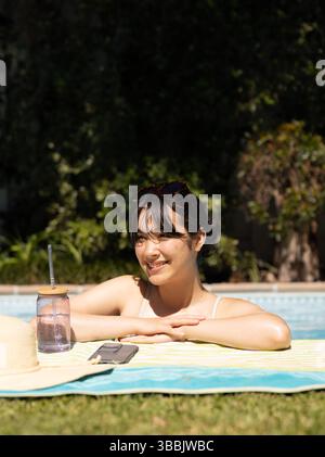 Entspannende asiatische Frau, die sich auf den Poolrand im Hinterhof lehnt, mit Strohhut-Smartphone-mason-Becher Stockfoto