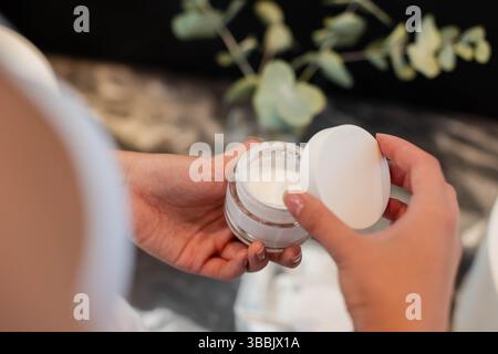 Asiatische Frau mit klarem Glascremebecher auf Marmorwaschtisch, mit Eukalyptuszweigen Stockfoto