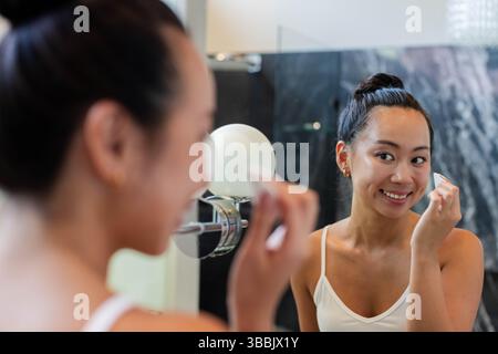Asiatische Frau, die Wattepad auf die Wange am Kosmetikspiegel im Badezimmer anbringt, mit runder Lampe, Marmorwand Stockfoto