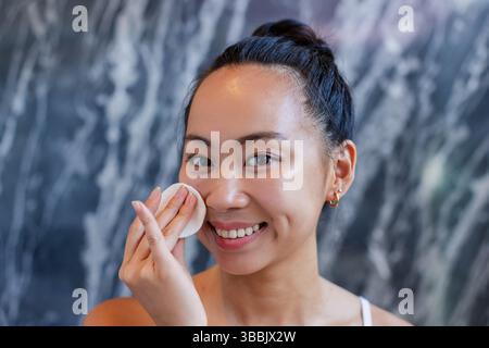 Lächelnde asiatische Frau, die im Badezimmer mit Marmorwand Baumwollkissen auf die Wange drückte, mit taufarbener Haut Stockfoto