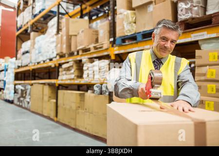 Reifer Erwachsener, der große Pappschachtel im Lagergang mit Bandspender, Kopierraum versiegelt Stockfoto