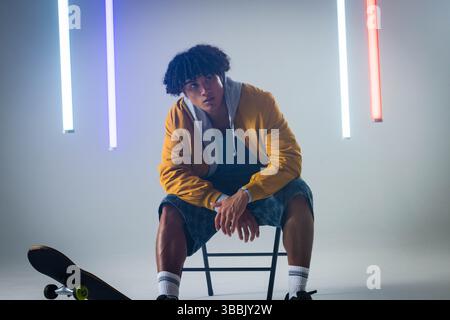 Posierender Mann mit Bomberjacke, der sich auf einen Klappstuhl im Lichtstudio stützt, mit Skateboard Stockfoto