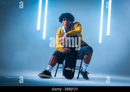 Skateboard halten, Afroamerikaner in seinen Zwanzigern auf Stuhl im Studio sitzen, mit LED-Röhren Stockfoto