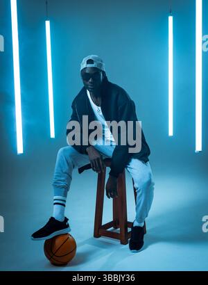 Afroamerikaner im Trainingsanzug sitzt im Studio auf dem Hocker und balanciert Basketball unter Neonröhren Stockfoto