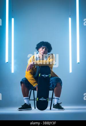 Sitzender Mann mit gelber Jacke auf Stuhl im Studio, hält Skateboard mit Neonröhrenlichtern Stockfoto