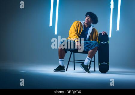 Sitzender afroamerikanischer Mann, der Jacke und Shorts auf dem Stuhl im Studio trägt und Skateboard hält Stockfoto