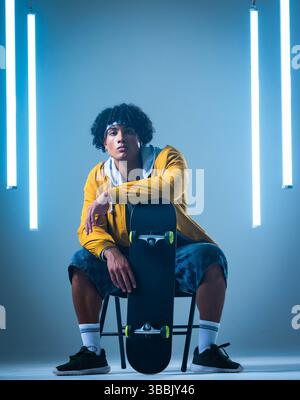 Sitzender afroamerikanischer Mann, der Skateboard senkrecht auf Stuhl im Studio hält, mit LED-Röhrenleuchten Stockfoto