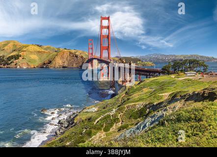 Die legendäre Golden Gate Bridge erstreckt sich über eine lebhafte blaue Bucht unter einem teilweise bewölkten Himmel mit üppigen grünen Hügeln, die das Wasser umgeben. Ein landschaftlich reizvolles Pat Stockfoto