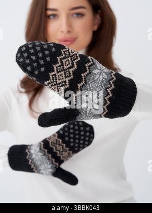 Gemütliche Fäustlinge mit nordischen Mustern in Schwarz, Grau und weiß werden von einer Frau in einem weißen Pullover präsentiert. Ihr Gesicht ist teilweise sichtbar und konzentriert sich auf Stockfoto