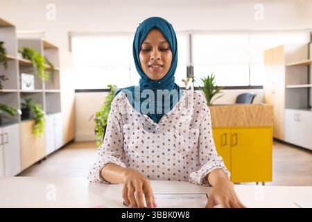 Junge Frau, die Dokumente an einem modernen Bürotisch mit Pflanzen und gelbem Schrank überprüft Stockfoto