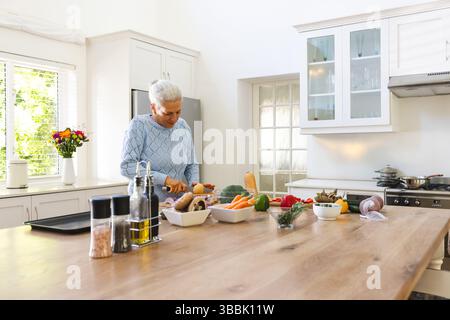 Ältere Frau, die Gemüse auf Holzinsel in der Küche hackt, mit Brett, Messer, Kopierraum Stockfoto