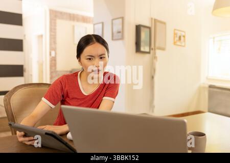 Junge asiatische Frau, die sich zu Hause an einen Holztisch lehnt, mit Laptop und Tablet mit Kaffeetasse Stockfoto