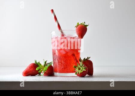 Erfrischendes Erdbeergetränk mit Eis in einer Glasschale, dekoriert mit einem Strohhalm und einer Scheibe Obst auf einem weißen Tisch mit Obst, Eis und Blättern mit einem Weißwein Stockfoto