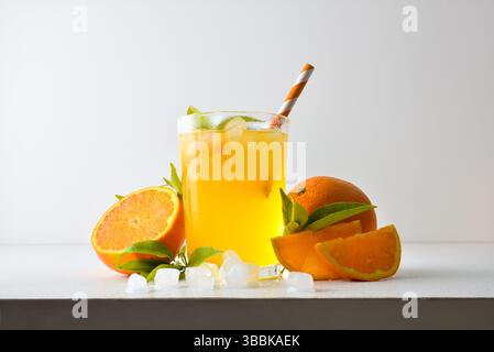 Erfrischendes Orangengetränk mit Eis in einer Glasschale, dekoriert mit einem Strohhalm und einer Scheibe Obst auf einem weißen Tisch mit Obst, Eis und Blättern mit einem weißen i Stockfoto
