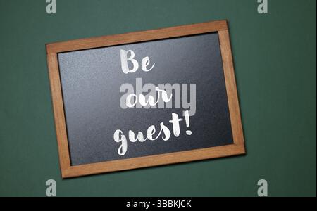 Kleine Tafel mit Phrase Be Our Guest auf dunkelgrünem Hintergrund, Draufsicht Stockfoto