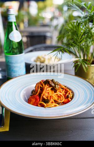 Ein Teller Spaghetti alla puttanesca, garniert mit grünen Oliven, Kapern und Kirschtomaten in einer reichhaltigen Tomatensoße. Das Gericht wird auf einem blauen Streifen serviert Stockfoto