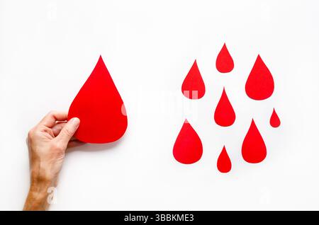 Eine weibliche Hand, die einen großen Papiertropfen Blut und ein paar kleinere Papiertropfen Blut auf weißem Hintergrund hält. Blutspendertagkonzept. Stockfoto