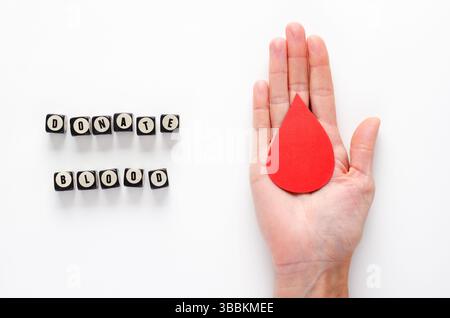 Eine weibliche Hand mit einem Papiertropfen Blut und einem Spendenbluttext in englischer Sprache, geschrieben mit Würfeln auf weißem Hintergrund. Blutspendertagkonzept. Stockfoto