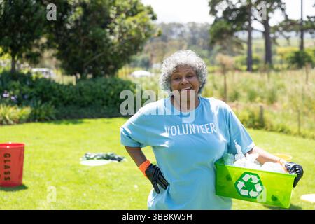Ältere Afroamerikanerin, die im Park ein freiwilliges Hemd trägt und einen grünen Recyclingbehälter hält Stockfoto